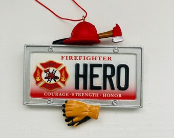 Può includere: Un ornamento decorativo con un elmetto da pompiere rosso, un'ascia e un design a targa. La targa recita "FIREFIGHTER HERO" con le parole "COURAGE, STRENGTH, HONOR" sottostanti. È inclusa anche un paio di guanti gialli e neri.