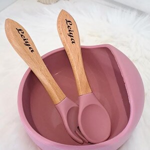 Peut inclure: Un bol en silicone rose avec deux cuillères en bois. Les cuillères sont gravées du nom "Leiya".