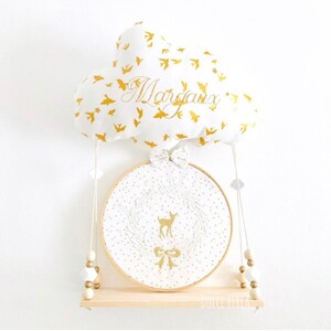 以下が含まれることがあります： 金色の鳥と「マルゴー」という名前が刺繍された白い雲の形をした枕。枕は、金色の水玉模様の白い輪と、その中に金色の鹿の刺繍が施された木製の棚から吊り下げられています。