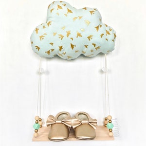 Peut inclure: Un coussin en forme de nuage bleu clair avec des silhouettes d'oiseaux dor&eacute;s est suspendu &agrave; une ficelle blanche au-dessus d'une &eacute;tag&egrave;re en bois avec des chaussures de b&eacute;b&eacute; dor&eacute;es et des perles en bois vertes et brunes. L'&eacute;tag&egrave;re porte le texte "Dolcetella".