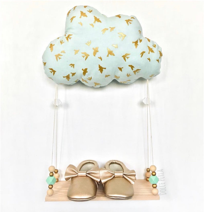 Peut inclure: Un coussin en forme de nuage bleu clair avec des silhouettes d'oiseaux dor&eacute;s est suspendu &agrave; une ficelle blanche au-dessus d'une &eacute;tag&egrave;re en bois avec des chaussures de b&eacute;b&eacute; dor&eacute;es et des perles en bois vertes et brunes. L'&eacute;tag&egrave;re porte le texte "Dolcetella".