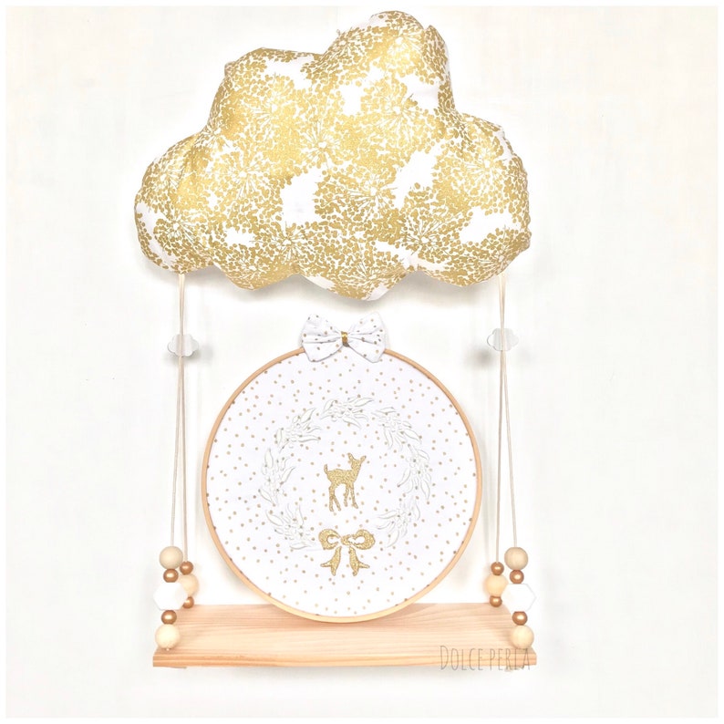 Peut inclure: Un nuage dor&eacute; &agrave; paillettes avec un n&oelig;ud blanc est suspendu au-dessus d'un cercle blanc et dor&eacute; avec un motif de cerf dor&eacute; &agrave; paillettes et de couronne. Le cercle est sur une &eacute;tag&egrave;re en bois avec des perles dor&eacute;es et blanches suspendues sur les c&ocirc;t&eacute;s. Le texte "DOLCE PERLA" est sur l'&eacute;tag&egrave;re.