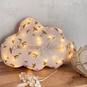 Peut inclure: Un coussin décoratif en forme de nuage, en tissu rose clair, orné de silhouettes d'oiseaux dorés et des noms "Juliana Adelina" en paillettes dorées. Le coussin est éclairé par des lumières intégrées, créant une lueur chaleureuse. Un arrangement floral décoratif est au premier plan.