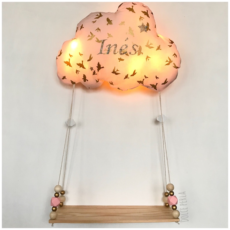 Peut inclure: Une veilleuse en forme de nuage rose avec des oiseaux dor&eacute;s et le nom "In&eacute;s" brod&eacute; dessus. La lumi&egrave;re est suspendue &agrave; une &eacute;tag&egrave;re en bois avec des perles roses et dor&eacute;es.