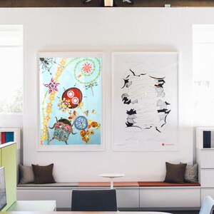 May include: Two framed pieces of art hang on a white wall in a modern office. The art on the left is a colourful abstract design with a blue background. The art on the right is a black and white line drawing. The office has a white bench with brown cushions and a desk with two chairs.