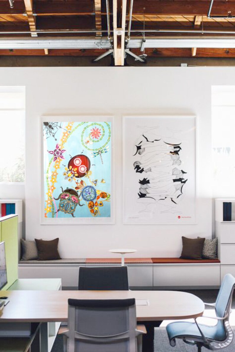May include: Two framed pieces of art hang on a white wall in a modern office. The art on the left is a colourful abstract design with a blue background. The art on the right is a black and white line drawing. The office has a white bench with brown cushions and a desk with two chairs.