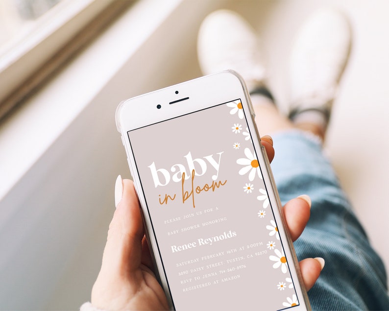 May include: A mobile phone screen displaying a baby shower invitation with a light pink background and white daisies. The invitation reads "baby in bloom" in white and orange text. The text also includes the name "Renee Reynolds", the date and time of the shower, and the address.