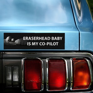 May include: A black bumper sticker with white text that reads "Eraserhead Baby Is My Co-Pilot". The sticker is on the back of a blue car.