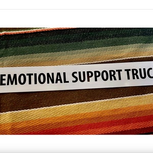 Puede incluir: Pegatina rectangular blanca con texto negro que dice "Emotional Support Truck". La pegatina está sobre un fondo de tela a rayas.