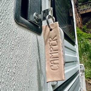 May include: A close-up of a silver key with a leather keychain that says "CAMPER" hanging in a door lock of a white camper van.