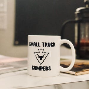 May include: White ceramic mug with black text that reads "Small Truck Campers" and a black silhouette of a bird with a bull's head inside a triangle.