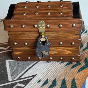 May include: A small wooden chest with a dark brown finish, decorated with brass studs and a black metal band. It is secured with a vintage-style padlock and has a brass clasp. The chest is a decorative storage item.
