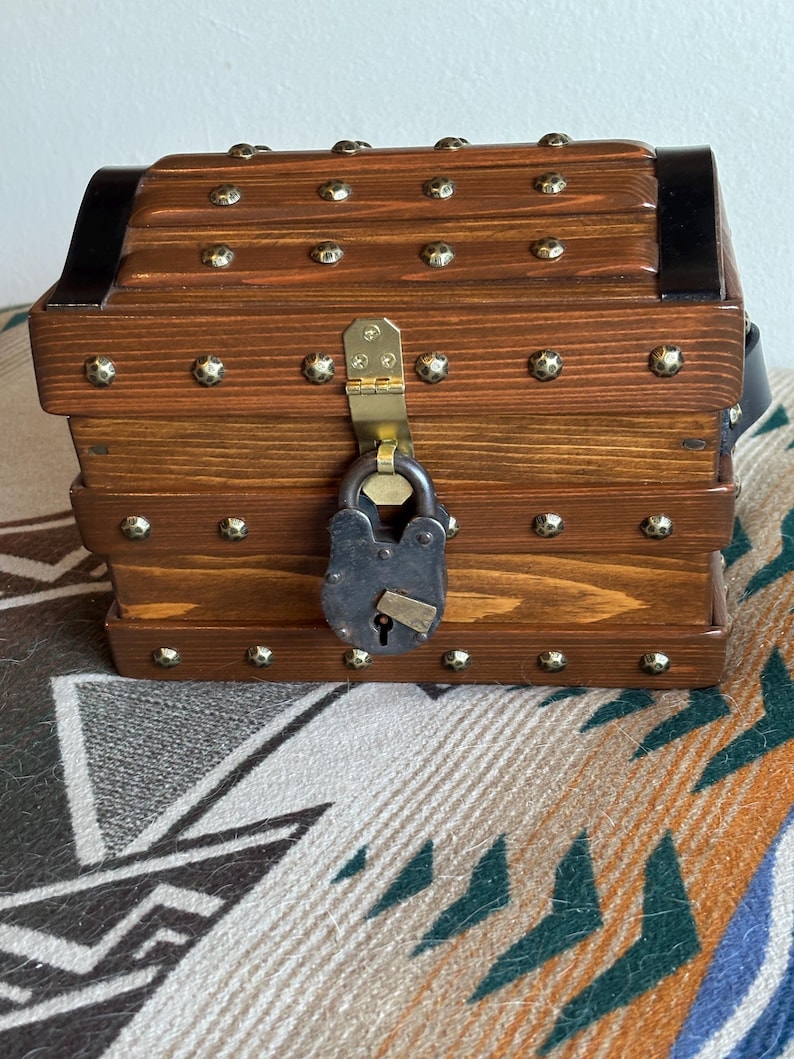 May include: A small wooden chest with a dark brown finish, decorated with brass studs and a black metal band. It is secured with a vintage-style padlock and has a brass clasp. The chest is a decorative storage item.