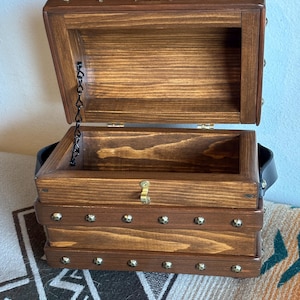 May include: A dark brown wooden treasure chest with an open hinged lid. The chest features brass-coloured studs and a metal clasp. A black leather handle is attached to the side. The interior is empty. The chest is reminiscent of a pirate's chest.