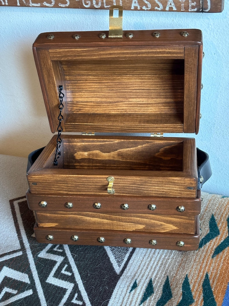May include: A dark brown wooden treasure chest with an open hinged lid. The chest features brass-coloured studs and a metal clasp. A black leather handle is attached to the side. The interior is empty. The chest is reminiscent of a pirate's chest.