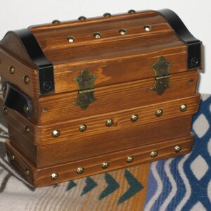 May include: A wooden chest with a curved top, black metal trim, and brass-coloured hardware. The chest is rectangular with rows of decorative studs. The wood has a warm, reddish-brown stain. A black handle is on the side.