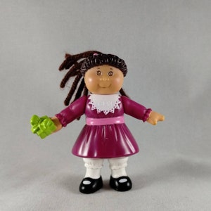 May include: A small plastic doll with dark brown yarn hair, wearing a magenta dress with a white collar featuring the text CPK, a pink waistband, white ruffled bloomers, and black shoes. The doll holds a small, bright green gift box in one hand.