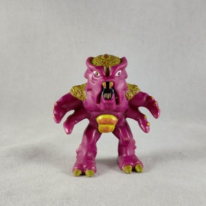 May include: A purple and green plastic toy monster with four arms and a toothy grin. The monster has a spiky head and a yellow and orange abdomen.