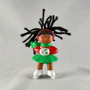 May include: A small plastic doll with dark skin and black yarn hair, wearing a red long-sleeved top, a green skirt, and white ice skates. The doll has red earmuffs and holds a white muff with a green holly leaf and red berry design.