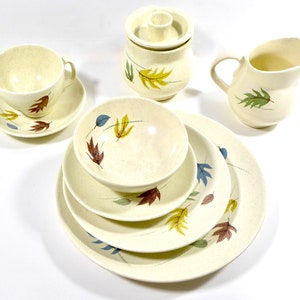 May include: Cream-colored vintage dish set with a speckled finish and leaf design. Includes a cup and saucer, sugar bowl, creamer, and various plates and bowls. The leaves are in shades of brown, blue, yellow, and green.