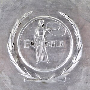 May include: Close-up of a silver-colored metal emblem. The emblem features a woman in a flowing gown holding a scale, surrounded by a laurel wreath. The word "EQUITABLE" is inscribed below the woman.