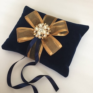 May include: A navy blue velvet pillow with a gold and white satin bow and a pearl and rhinestone brooch. The pillow is tied with a navy blue ribbon.