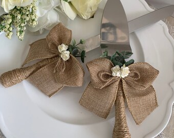 May include: A set of two cake servers with burlap bows and white flowers. The servers are on a white plate.
