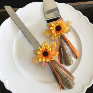May include: A set of two cake cutting knives decorated with sunflowers and brown and orange ribbon. The knives are on a white plate.