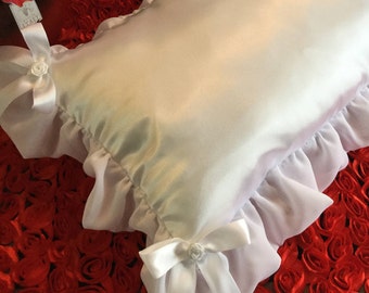 May include: White satin pillow with a ruffled edge and white satin bows with a small white rose accent. The pillow is sitting on a red rose background.