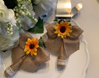May include: A cake knife and server set with burlap bows and sunflower accents. The handles are wrapped in twine and finished with lace. The set is displayed on a white plate, perfect for a rustic wedding.