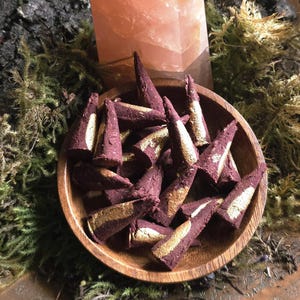 May include: A pink quartz crystal point is behind a wooden bowl filled with maroon incense cones with gold detailing. The bowl rests on moss and bark.
