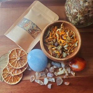 May include: A wooden bowl filled with dried herbs and flowers, including yellow petals, green leaves, and brown seeds. A blue crystal sphere, a small orange crystal, and a brown paper bag with dried herbs and flowers are also visible on a wooden surface.