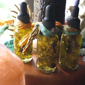 May include: Three clear glass dropper bottles filled with a yellow-tinted liquid containing visible botanical pieces. Each bottle features a black dropper cap and is tied around the neck with a strip of frayed fabric in shades of orange or green.