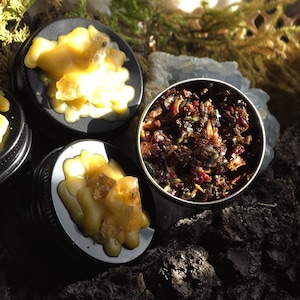 Puede incluir: Primer plano de tres pequeños recipientes con tapa negra. Dos contienen sustancias cerosas amarillas con inclusiones cristalinas. El tercer recipiente contiene una mezcla de hierbas y flores secas. El fondo presenta musgo y corteza.