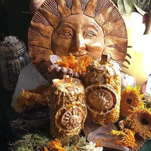 Op de afbeelding: Een decoratieve opstelling met een zon-gezicht plaque en twee cilindrische kaarsen. De kaarsen zijn versierd met zon-medaillons en gedroogde bloemen. De scène wordt geaccentueerd met gedroogde zonnebloemen, mos en kristallen, wat een warme, aardse esthetiek creëert.