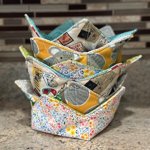 Puede incluir: Un juego de tres protectores de cuenco de tela, apilados uno encima del otro. El protector de cuenco inferior es blanco con un patrón floral, el protector de cuenco del medio es amarillo con un patrón de oso koala, y el protector de cuenco superior es azul con un patrón de póster de viaje vintage.