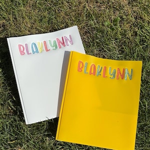 May include: Two notebooks on grass. One is white, the other is yellow. Both have the name "BLAKLYNN" embroidered in colorful thread across the top. The notebooks are open, and the grass is green.