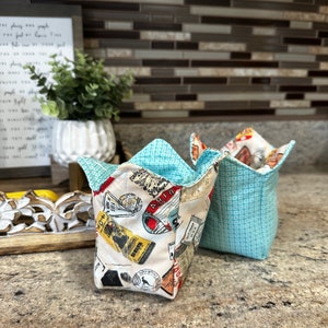 Puede incluir: Dos cestas de almacenamiento de tela, una con un patrón geométrico azul y blanco y la otra con un estampado vintage de temática de viaje. Las cestas están sobre una encimera de color marrón claro.