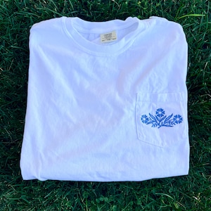 Puede incluir: Una camiseta blanca con un bolsillo con un diseño floral azul. La camiseta está doblada sobre hierba verde. Tiene cuello redondo y mangas cortas.