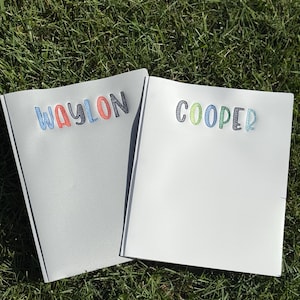 以下が含まれることがあります： 刺繍された名前が書かれた2冊のノート。1冊はグレーで、青、赤、オレンジ色の糸で「WAYLON」と書かれています。もう1冊は白で、緑、青、グレーの糸で「COOPER」と書かれています。ノートは芝生の上にあります。