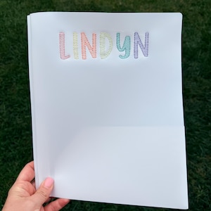 May include: A white plastic folder featuring the name LINDYN embroidered in pastel-colored thread across the top. The letters are stitched in a zigzag pattern using light pink, orange, yellow, light green, blue, and purple thread colors.