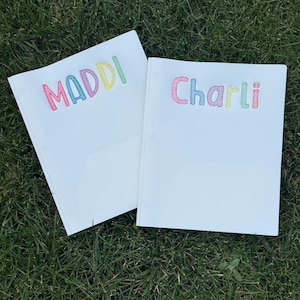 May include: Two white notebooks with the names "Maddi" and "Charli" embroidered in colourful thread. The notebooks are lying on a bed of green grass.