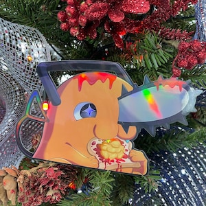 May include: A Christmas tree bauble depicting a cartoon character with orange skin, a blue eye, and a chainsaw for a nose. The character is eating a slice of toast with peanut butter and jelly.