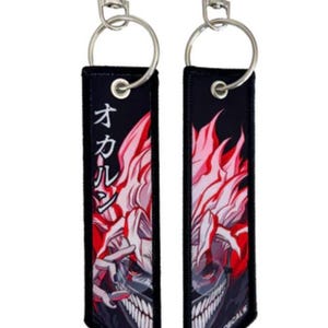 Può includere: Due portachiavi neri con anelli e clip argentati. Ogni portachiavi presenta un personaggio anime rosso e bianco con testo giapponese che recita "オカル" su un portachiavi. Il testo "MODIFIED DECALS" è stampato sul fondo di un portachiavi.