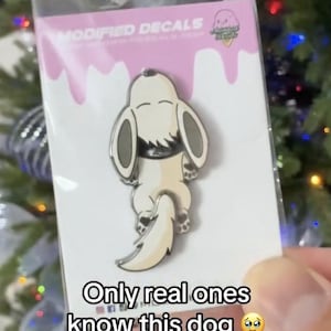 May include: A pin featuring a cartoon dog in a lying down pose, with a cream-colored body and a white tail. The pin is on a pink and white backing with the text "Only real ones know this dog". The brand name "Modified Decals" is visible.