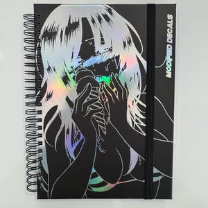 Puede incluir: Cuaderno negro con espiral con una ilustración holográfica de una persona. La ilustración presenta un contorno negro con acentos holográficos. El texto "MODIFIED DECALS" está impreso verticalmente en el lado derecho del cuaderno.