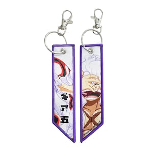 以下が含まれることがあります： 紫色の縁取りが付いた2つの白いキーホルダー。それぞれにアニメキャラクターのイラストが描かれています。1つは日本語のテキストと赤と白のデザインです。もう1つは「MODIFIED DECALS」というテキストが入った笑顔のキャラクターです。