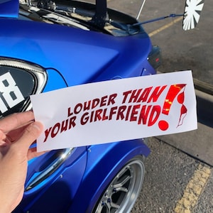 Puede incluir: Una pegatina rectangular blanca con el texto "LOUDER THAN YOUR GIRLFRIEND!" en rojo, con un gráfico de sujetador rojo. La pegatina se sostiene frente a un coche azul.