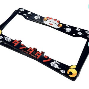 May include: Black license plate frame with a Japanese lucky cat design. The frame features red Japanese text, white clouds, and gold accents. The lucky cat has a white body, red collar, and is waving its paw.