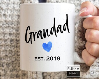 Puede incluir: Taza de cerámica blanca con texto negro que dice "Grandad Est. 2019" y un corazón azul acuarela.